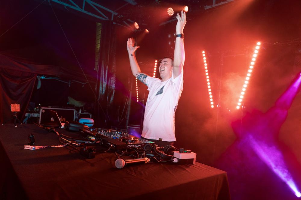 DJ con los brazos en alto detrás de la mesa de mezclas, con luces rojas y humo.