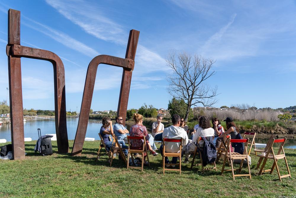 Grupo de personas conversando sentadas en círculo junto a una escultura de hierro y la ría, en la Feria Ribera.
